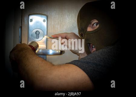 Un homme masqué brillant une lampe de poche sur une porte qu'il essaie d'ouvrir avec des outils Banque D'Images