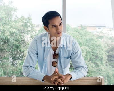Portrait d'un homme asiatique attentionné qui se sent seul à regarder loin et à penser dans la chambre à la maison près de la fenêtre en verre le matin. Banque D'Images