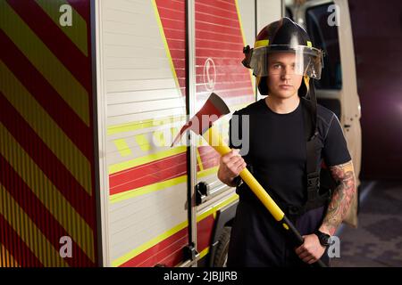 Photo d'un pompier portant un casque avec une hache contre un moteur d'incendie. Banque D'Images