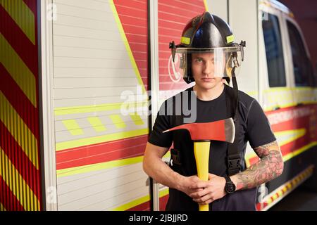 Photo d'un pompier portant un casque avec une hache contre un moteur d'incendie. Banque D'Images