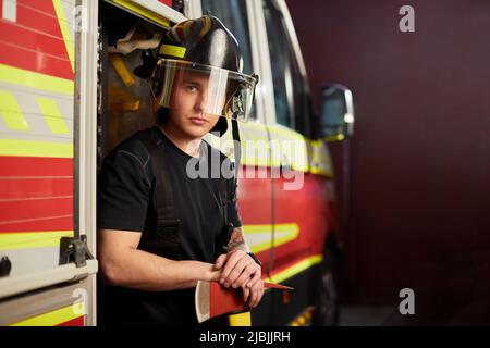 Photo d'un pompier portant un casque avec une hache contre un moteur d'incendie. Banque D'Images