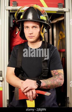 Photo d'un pompier portant un casque avec une hache contre un moteur d'incendie. Banque D'Images