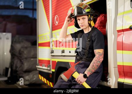 Photo d'un pompier portant un casque avec une hache contre un moteur d'incendie. Banque D'Images