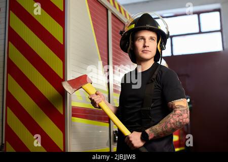 Photo d'un pompier portant un casque avec une hache contre un moteur d'incendie. Banque D'Images