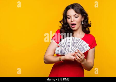 Agréablement surpris femme d'affaires porte rouge regardant le ventilateur de l'argent liquide dans des billets en dollars dans fond jaune en studio. Affaires, finance, savi Banque D'Images