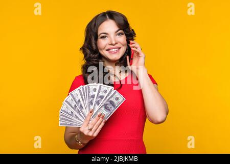 Business Woman détient un fan de billets en dollars et parle sur cellule en fond jaune. Fille financièrement savvy utilisant le smartphone. Finance, investissement Banque D'Images