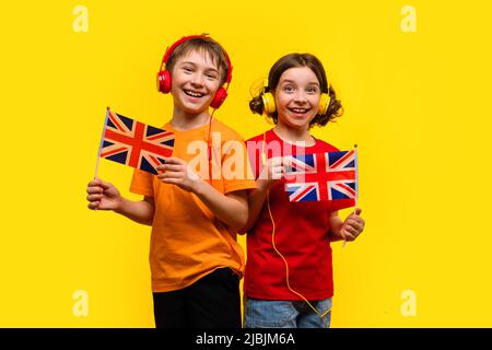 Deux enfants de l'école avec casque peuvent accueillir des drapeaux britanniques. Happy Smart Brother et Sister 9-11 y.o en T-shirts Bright Basic posant sur fond jaune. Con Banque D'Images