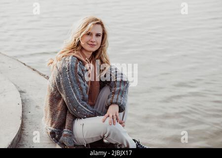 Bonne femme blonde souriante dans des vêtements chauds assis sur la rive de la rivière seul le jour frais ensoleillé, relaxant, méditant et appréciant la paix et la liberté Banque D'Images