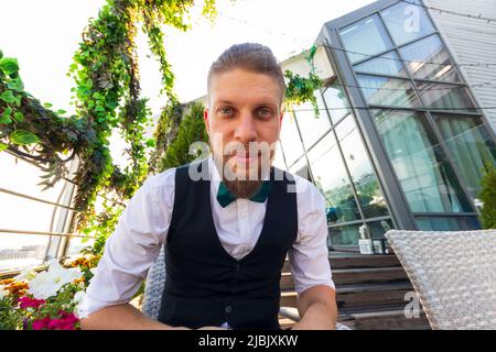 Jeune homme barbu dans une chemise blanche, gilet noir et noeud papillon regarde la caméra tout en étant assis dans un penthouse Banque D'Images