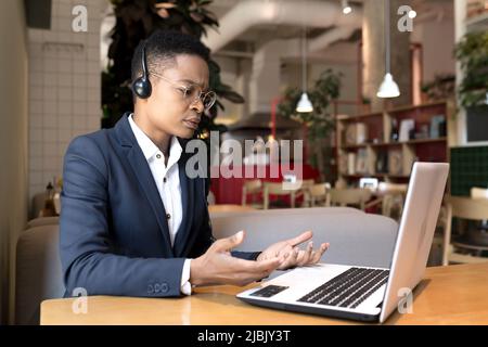 Une femme afro-américaine sérieuse utilise un micro-casque pour un appel vidéo, travaille dans un café, pendant un voyage d'affaires touristique, une femme d'affaires concentrée et réfléchie Banque D'Images