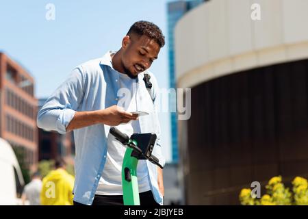 Location d'un scooter électrique. Jeune afro-américain louant un transport écologique, payant en ligne avec un téléphone portable Banque D'Images