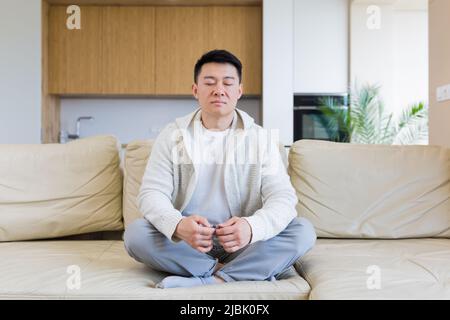 le jeune homme asiatique est assis sur un canapé en position lotus et médite dans le don Banque D'Images