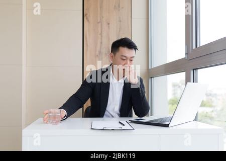 un jeune travailleur de bureau asiatique souffrant de maux de tête au travail boit de l'eau une pilule de médicament. Homme malade dans un costume à l'ordinateur à l'intérieur wi Banque D'Images