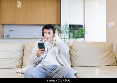 jeune homme asiatique joyeux écoutant de la musique dans un casque installé sur un canapé à la maison. Un homme seul sur le canapé se sent un bon style de vie. Heureux homme avec le Banque D'Images