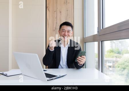 Joyeux jeune homme d'affaires asiatique au bureau, regardant la caméra, avec des émotions de gagnant ou de victoire, la promotion financière Paris sportifs. L'homme s'exclame Banque D'Images