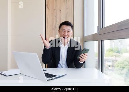 Joyeux jeune homme d'affaires asiatique au bureau, regardant la caméra, avec des émotions de gagnant ou de victoire, la promotion financière Paris sportifs. L'homme s'exclame Banque D'Images