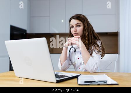 Jeune femme médecin traite en ligne de la maison, consulte par un appel vidéo à partir d'un ordinateur portable, assis dans la cuisine dans un manteau blanc, regardant le chamer Banque D'Images