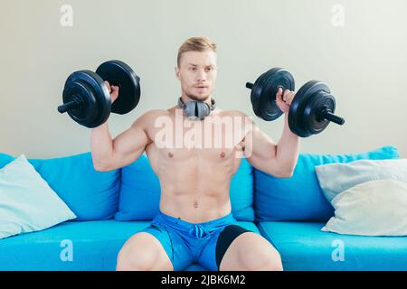 Un jeune homme sportif élégant s'entraîne avec des haltères, fait du sport à la maison, s'assoit sur un canapé, écoute de la musique sur un casque, regarde la caméra Banque D'Images