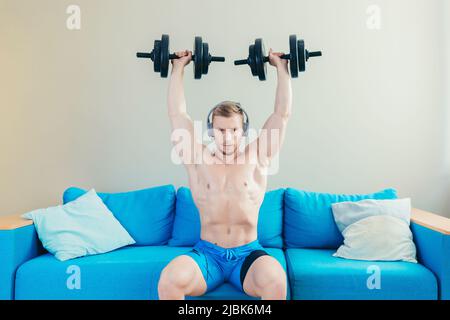 Un jeune homme sportif élégant s'entraîne avec des haltères, fait du sport à la maison, s'assoit sur un canapé, écoute de la musique sur un casque, regarde la caméra Banque D'Images