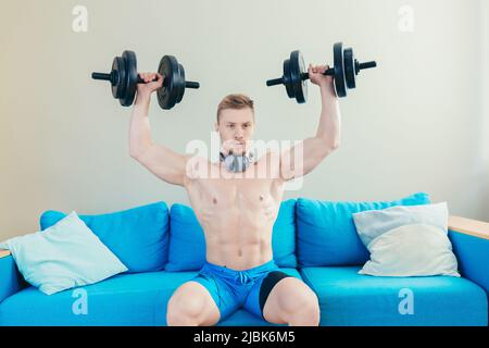 Un jeune homme sportif élégant s'entraîne avec des haltères, fait du sport à la maison, s'assoit sur un canapé, écoute de la musique sur un casque, regarde la caméra Banque D'Images