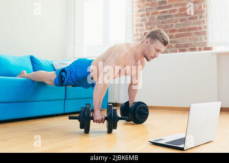 Jeune homme sportif élégant entraîneur s'entraîne en ligne avec ordinateur portable, faisant des exercices à la maison sur un tapis avec des haltères Banque D'Images
