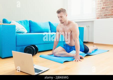 Jeune homme sportif élégant entraîneur s'entraîne en ligne avec ordinateur portable, faisant des exercices à la maison sur un tapis avec des haltères Banque D'Images