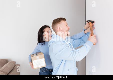 Jeune couple, homme et femme font des réparations ensemble dans un nouvel appartement, maison, homme clous une image au mur Banque D'Images