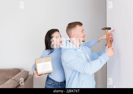 Jeune couple, homme et femme font des réparations ensemble dans un nouvel appartement, maison, homme clous une image au mur Banque D'Images