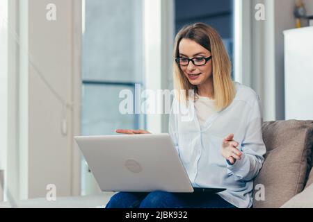 Belle femme blonde dans des vêtements décontractés assis sur un canapé à la maison, et utilise un ordinateur portable pour la communication vidéo avec les amis et la famille Banque D'Images