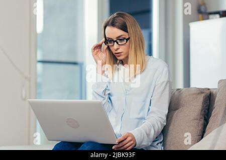 Belle femme blonde dans des vêtements décontractés assis sur un canapé à la maison, et utilise un ordinateur portable pour la communication vidéo avec les amis et la famille Banque D'Images