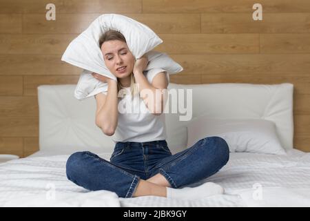 Une jeune femme en colère assise sur le lit à la maison, dans une chambre d'hôtel, couvre ses oreilles avec un oreiller provenant du bruit fort des voisins, des réparations, du bruit de la Banque D'Images