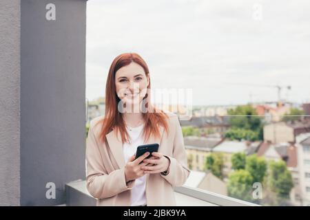 Jeune belle femme correspondait au téléphone, souriant et habillé dans un costume d'affaires debout sur le balcon Banque D'Images