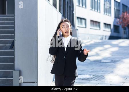 Belle femme asiatique parlant au téléphone, femme d'affaires sur une pause à pied près du bureau Banque D'Images