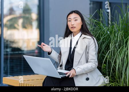 Femme d'affaires asiatique prospère travaillant sur un ordinateur portable à l'extérieur, assis près d'un bureau et regardant l'appareil photo Banque D'Images