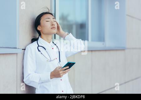 Triste médecin infirmière asiatique, près de la clinique pendant une pause, utilise le téléphone, la femme a un mal de tête sévère, déprimé surtravailleur Banque D'Images