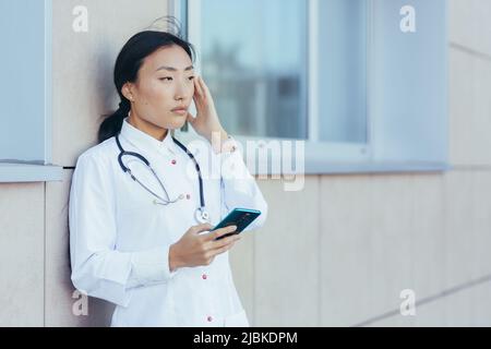 Triste médecin infirmière asiatique, près de la clinique pendant une pause, utilise le téléphone, la femme a un mal de tête sévère, déprimé surtravailleur Banque D'Images