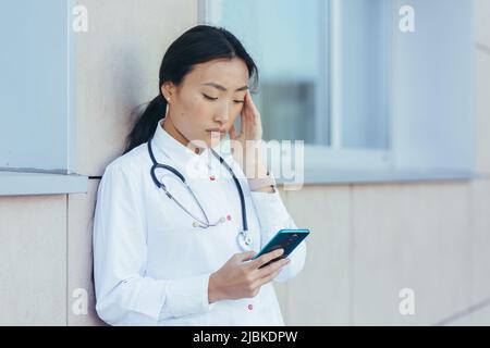 Triste médecin infirmière asiatique, près de la clinique pendant une pause, utilise le téléphone, la femme a un mal de tête sévère, déprimé surtravailleur Banque D'Images