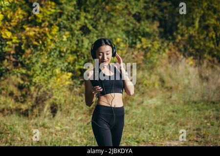 Jeune femme asiatique belle, lors d'une promenade matinale dans le parc, l'athlète court, et écoute de la musique dans les grands écouteurs utilise le téléphone, et une application pour Banque D'Images