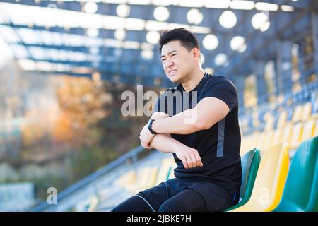 L'athlète a une douleur sévère dans son bras, l'homme asiatique s'assoit sur une chaise dans le stade, s'agenouillent son bras, blessé pendant l'entraînement le matin à l'extérieur, se reposant Banque D'Images