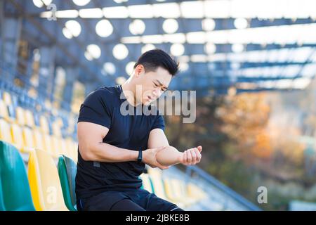 L'athlète a une douleur sévère dans son bras, l'homme asiatique s'assoit sur une chaise dans le stade, s'agenouillent son bras, blessé pendant l'entraînement le matin à l'extérieur, se reposant Banque D'Images
