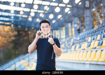 Athlète asiatique réussi parlant au téléphone, partageant ses réalisations et ses résultats sportifs, souriant et se réjouissant, à l'extérieur du sport stadiu Banque D'Images