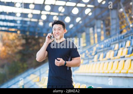 Athlète asiatique réussi parlant au téléphone, partageant ses réalisations et ses résultats sportifs, souriant et se réjouissant, à l'extérieur du sport stadiu Banque D'Images