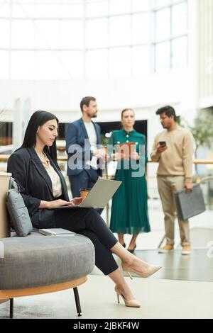 Portrait complet d'une femme d'affaires élégante du Moyen-Orient utilisant un ordinateur portable tout en étant assise dans la salle d'attente du bâtiment de bureau Banque D'Images