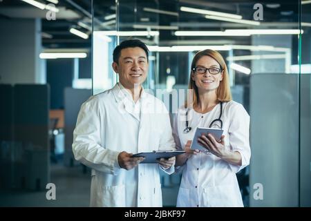 Portrait de deux médecins, équipe d'homme asiatique et de femme caucasienne regardant la caméra et souriant, travaillant dans un bureau de clinique moderne Banque D'Images