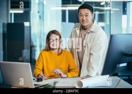 Équipe créative d'architectes designers travaillant sur un projet à une table dans un bureau moderne, asiatique homme et femme regardant la caméra et souriant, bâtiment Banque D'Images