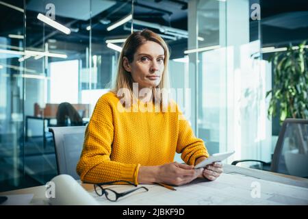 Femme architecte travaille avec des dessins sur le bureau, regarde sérieusement la caméra, utilise une tablette Banque D'Images