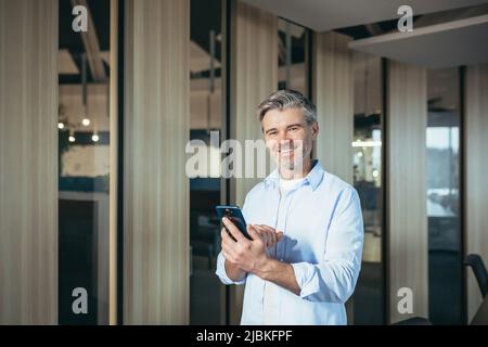 Homme réussi utilise une application mobile sur téléphone pour les rencontres, homme d'affaires expérimenté et aux cheveux gris au bureau Banque D'Images