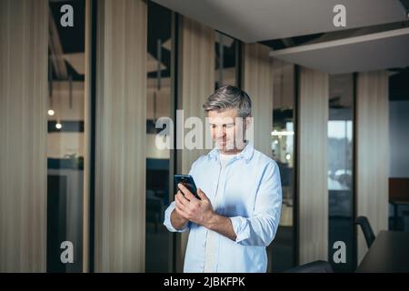 Homme réussi utilise une application mobile sur téléphone pour les rencontres, homme d'affaires expérimenté et aux cheveux gris au bureau Banque D'Images