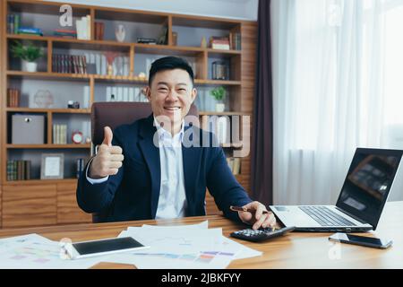 Portrait d'un financier asiatique, d'un homme d'affaires travaillant au bureau, de la paperasserie, regardant l'appareil photo et souriant, montrant le pouce vers le haut Banque D'Images