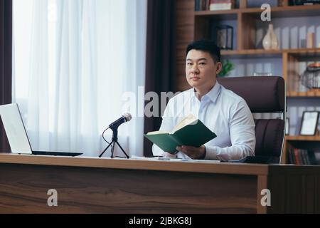 Un prédicateur asiatique lit la bible en ligne, enregistre un podcast, s'assoit à une table et regarde la caméra, utilise un microphone, tient sa main dans son cœur, onl Banque D'Images
